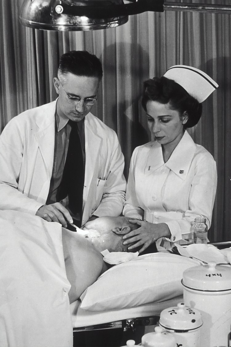 A Caucasian physician is changing the dressing on a patient's neck, while a Caucasian nurse holds the patient's head. Photo was taken at Public Health Service Tumor Clinic, Marine Hospital, Baltimore. 1950